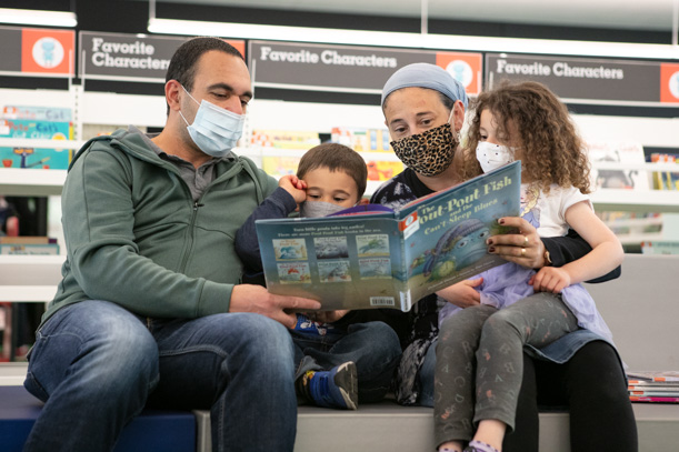 A family of four, with two children and two parents, all wearing masks, sit together to read a picture book.