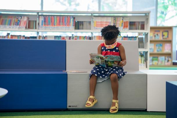 A young girl sits on a silver and blue couch, reading a book. She has a blue dress with white stars and yellow sandals.