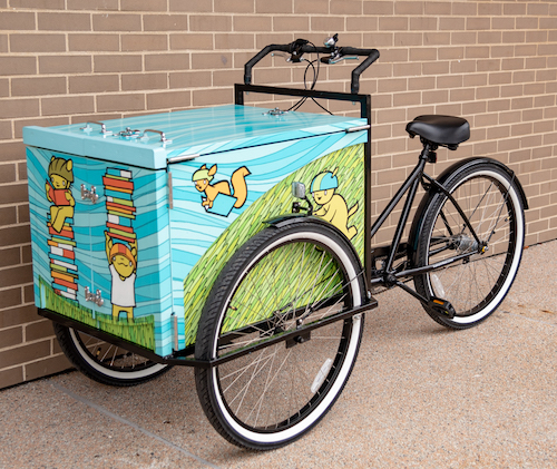 The book bike looks like a tricycle with the double wheels in the front, flanking a blue and green painted box with characters holding books and wearing helmets.
