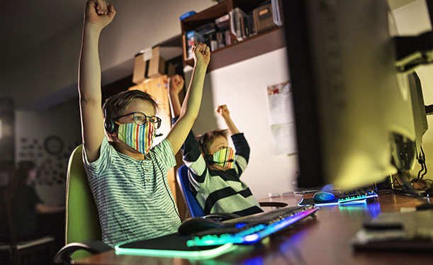 Two kids wearing masks sit at a desk with computer devices in front of them and arms raise overheard triumphantly.