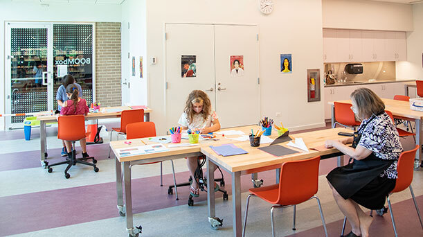 Three children and a Skokie Public Library staff member, all masked, sit in orange chairs inside the BOOMbox, which has posters on the wall, activities on the tables, and a glass door in the back. 