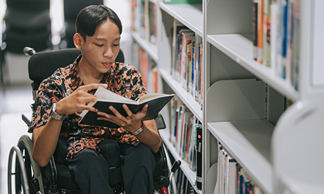 a person using a wheelchair is looking down at a book; they are next to a library bookshelf