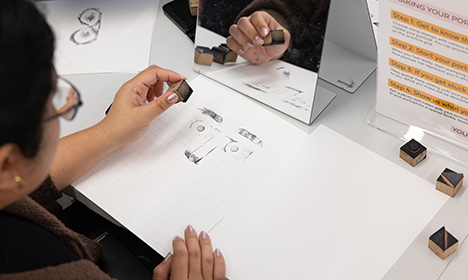 top down video of a woman holding shape stamps while looking at herself in a small mirror. On the table in front of her is a portrait made out of stamps