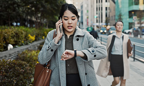 woman walking on a sidewalk. A cellphone is to her ear and she looks down at the watch on her wrist