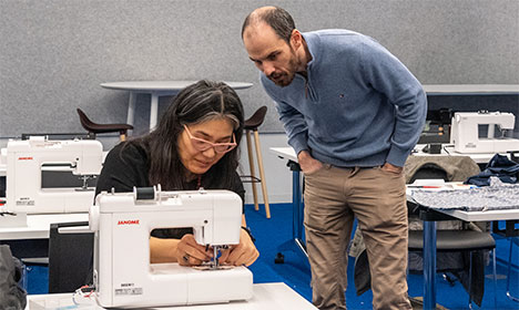 a woman is seated while working on a sewing machine while a man stands over her and watches
