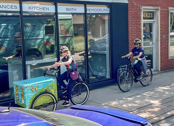 A woman rides the book bike on a sidewalk and she is giving the thumbs up. Another woman rides behind her on a regular bike. The book bike has a large blue and green box between the two front wheels, which holds books.