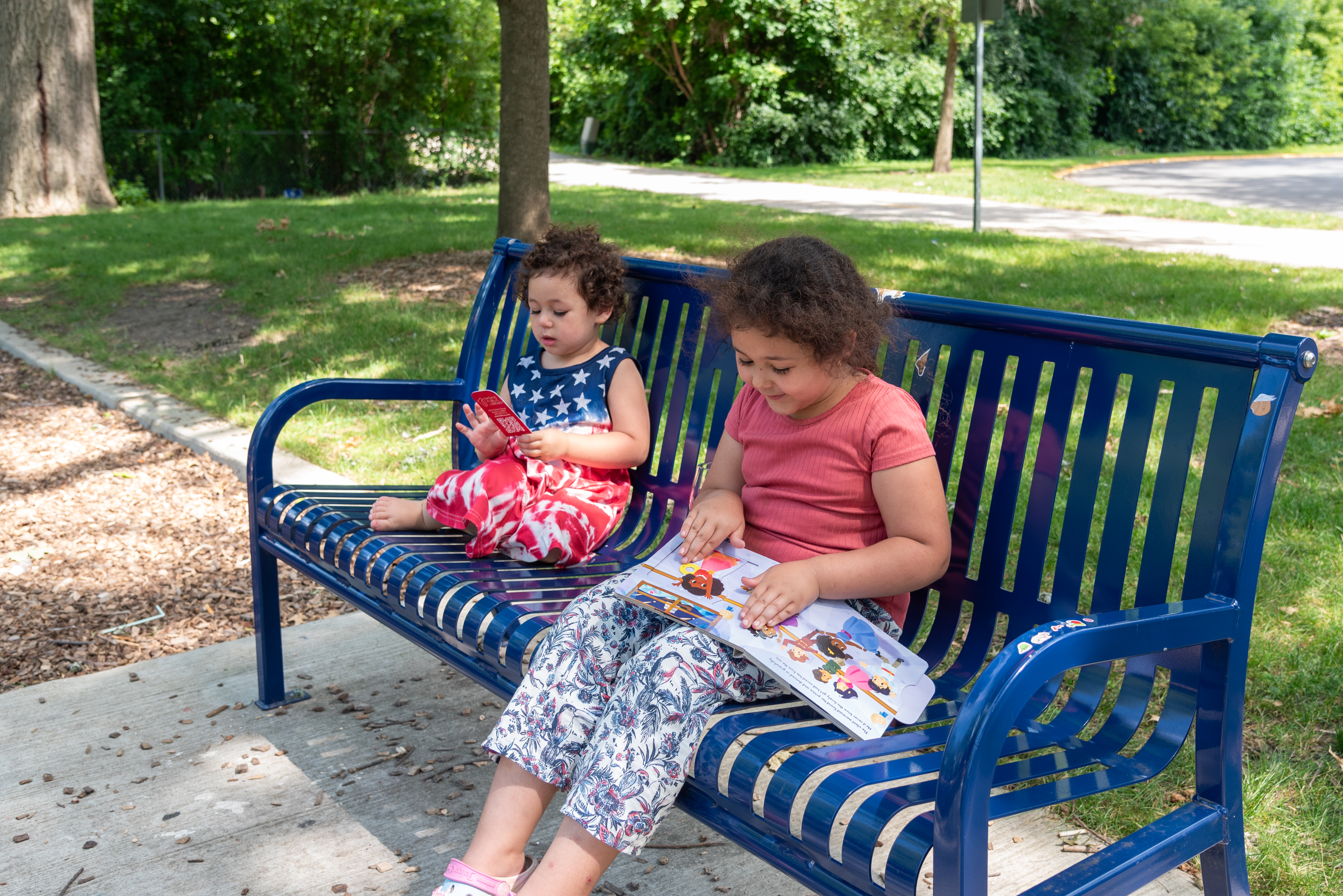 Two young children sitting on a park bench. One is playing with a bookmark and the other is reading a picture book. 