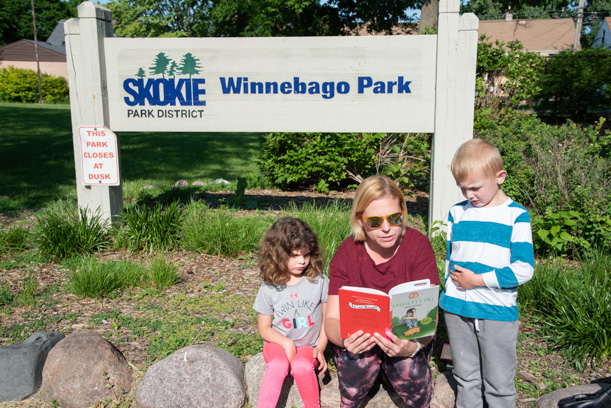 A woman in a red shirt reads to two children, one boy wearing a blue and white striped shirt and one girl wearing a gray shirt and pink pants. There is a sign behind where they sit, which reads "Winnebago Park" and has a logo for the Skokie Park District.