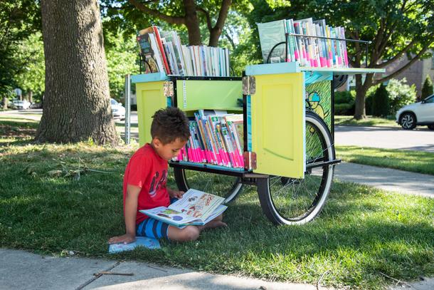 A child in a red t-shirt sits in the grass reading a picture book in front of the Skokie Public Library book bike, which is a yellow and blue open box on wheels, filled with books.