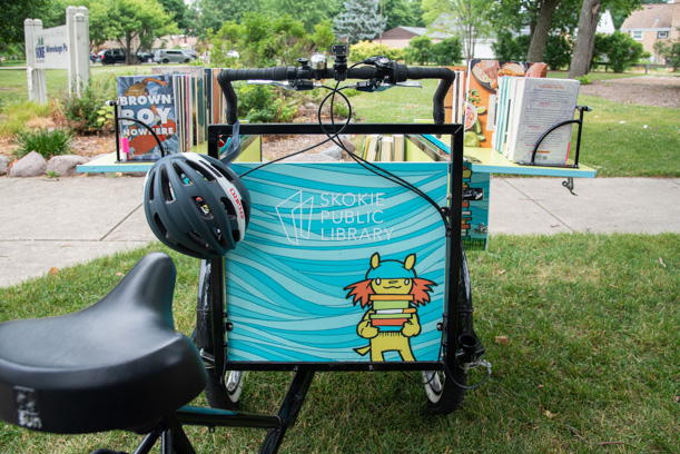 A black bike seat in the left corner leads to the back of a cargo hold on the Skokie Public Library book bike, which is blue with an animal character holding books painted on the back. There is a gray helmet hanging from the handle bars and the cargo hold is open, revealing shelves that hold two rows of books.