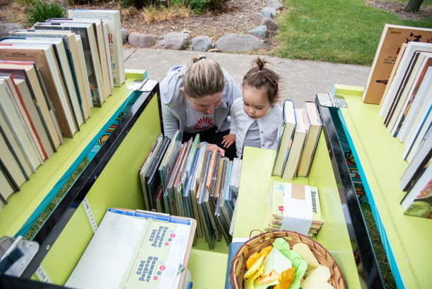 A woman an a very young girl look at books in the cargo hold of the Skokie Public Library book bike. The hold is yellowish green and filled with books, summer reading pamphlets, and a brown basket of paper origami hearts.