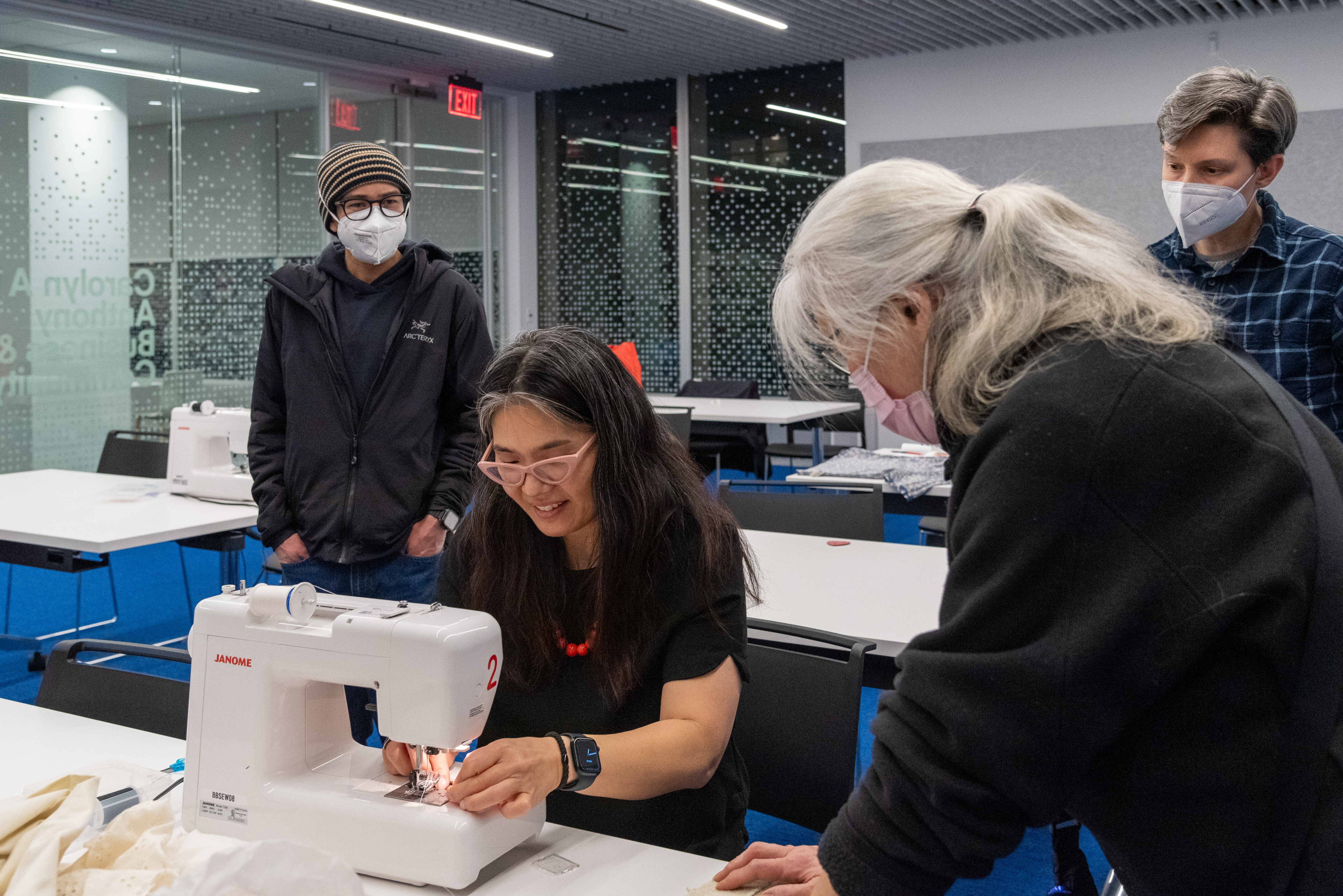 Julie is sitting before a sewing machine while three adults gather around her. Julie is mending a garment. Julie is sitting before a sewing machine while three adults gather around her. Julie is mending a garment.