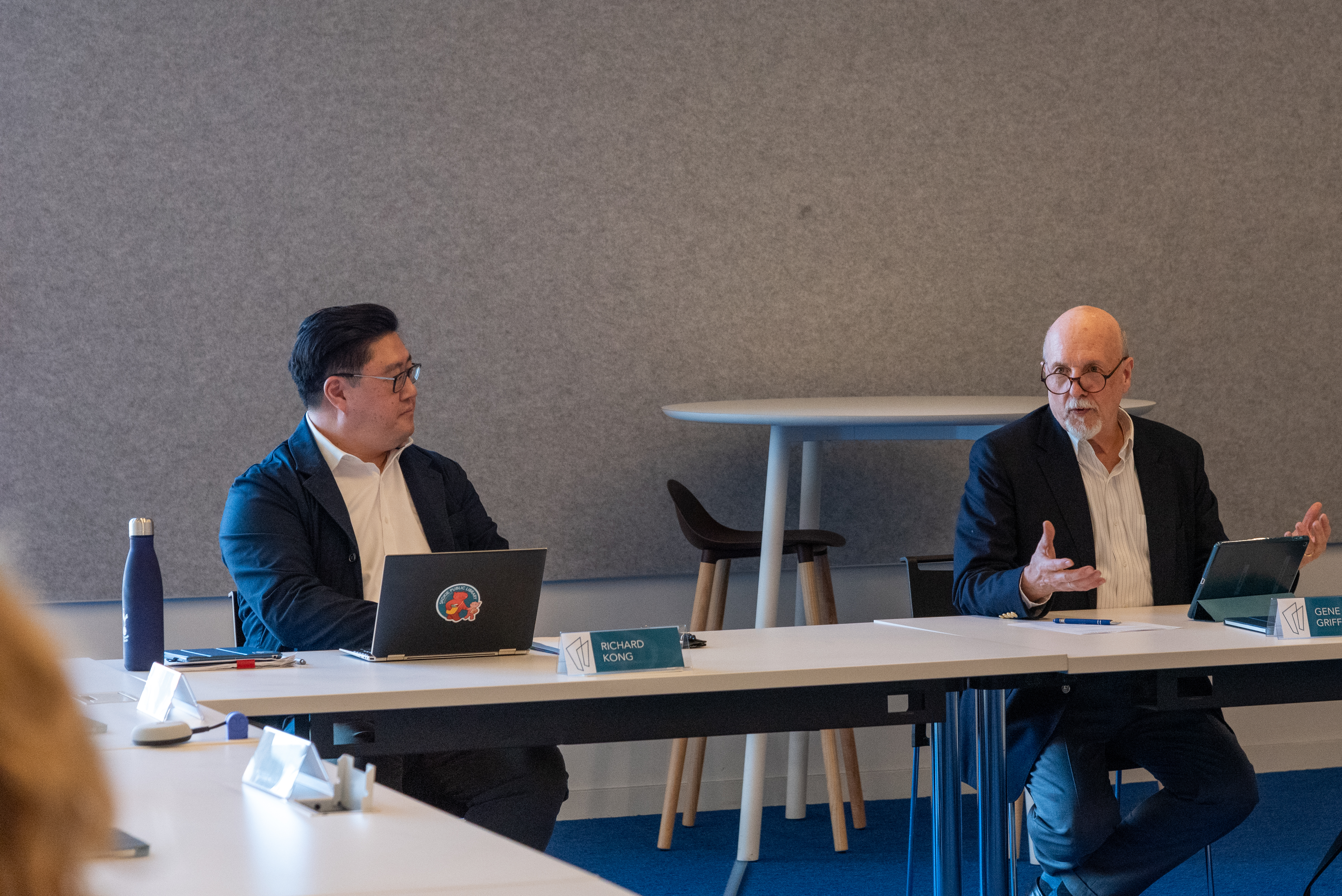 Two men seated at a table at the head of the room. The man on the right is speaking and gesturing. Two men seated at a table at the head of the room. The man on the right is speaking and gesturing.