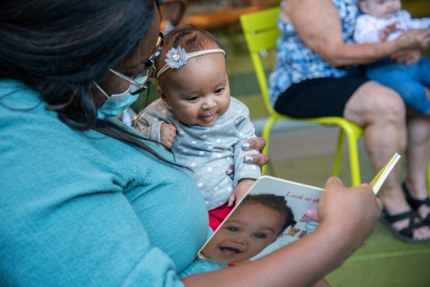 A mother reads to her baby, who is wearing a flower headband and smiling, from a board book about facial expressions.