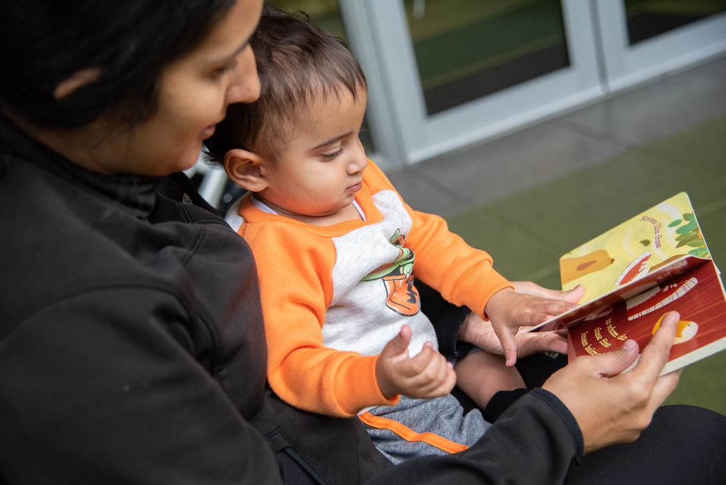 A woman reads a board book to a child seated on her lap