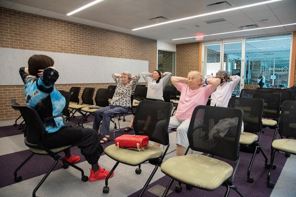 Four older adults hold their hands behind their head as part of an exercise class. An instructor in a blue shirt sits in front of them, leading the exercise.