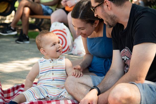 A toddler wearing a striped sleeveless jumper looks up at their parents with a smile. The parents, sitting on a red and white blanket, are smiling back.