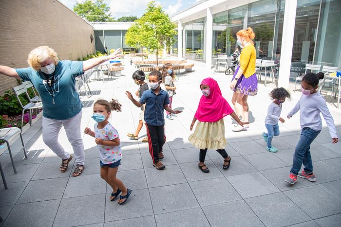 A group of young children dance along with library staff outside in the courtyard.