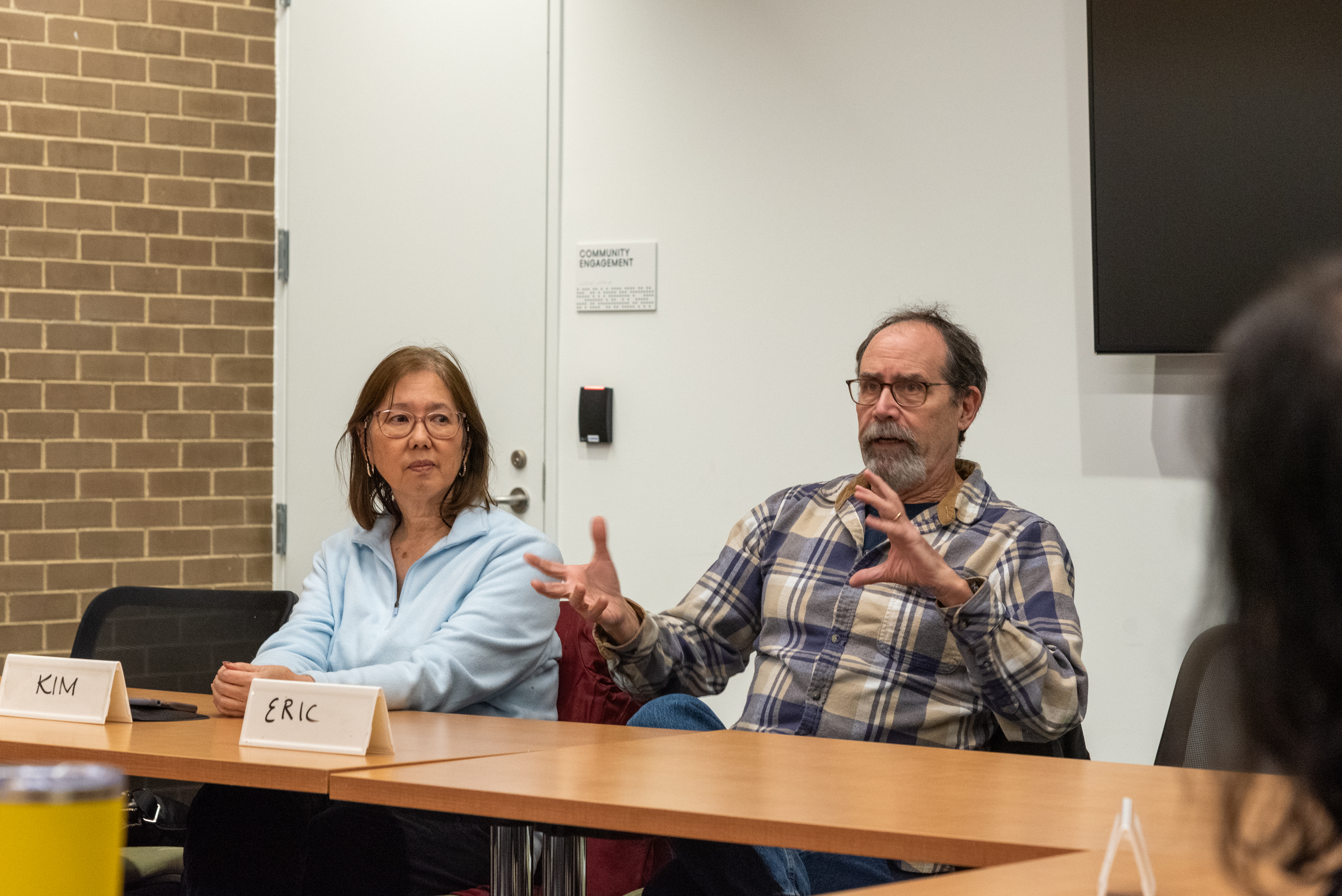 Two people sitting at a table next to each other. One person is gesturing and speaking Two people sitting at a table next to each other. One person is gesturing and speaking
