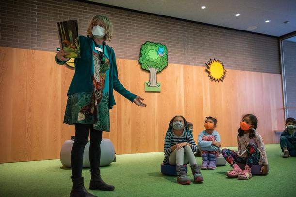 A librarian with blonde hair and wearing a green jacket stands and reads from a picture book as three children sit on the floor and listen. There is a tree as well as a sun on the wall behind them.