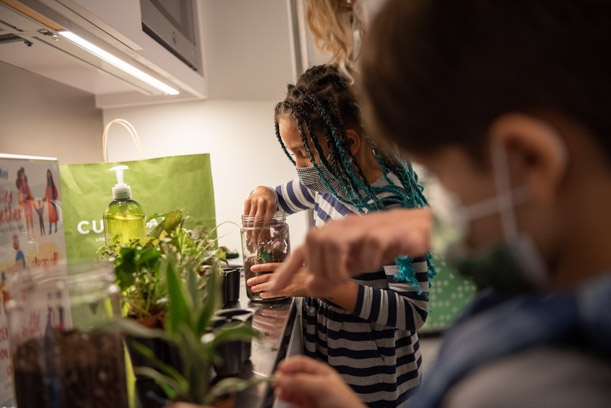A young patron with long blue and black hair puts her hand into a glass jar full of dirt. There are small plants and another small patron in the foreground.