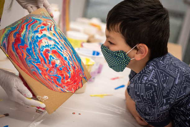 A young boy with a green polka-dot mask looks intently at a swirled red, blue, and yellow painting being held up by two gloved hands.