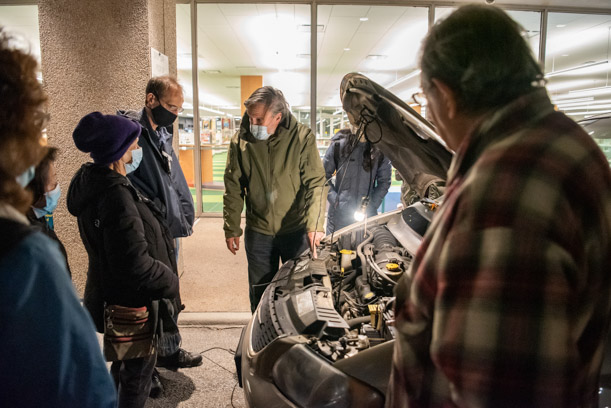 Patrons surround the open hood of a car, listening to an instructor who is pointing to something under the hood. There is a light bulb illuminating what's under the hood, as it is nighttime.