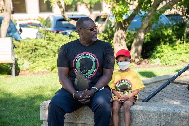 An adult and a child sit on a stone ledge on the Village Green in downtown Skokie. They are both wearing t-shirts with the silhouette of a woman with an afro and text about the Skokie Juneteenth Celebration.
