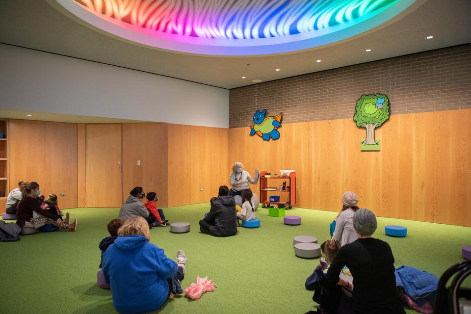 A staff person reads from a picture book to families in the Storytime Room, which features colorful characters and a ceiling mural by artist Jay Ryan.