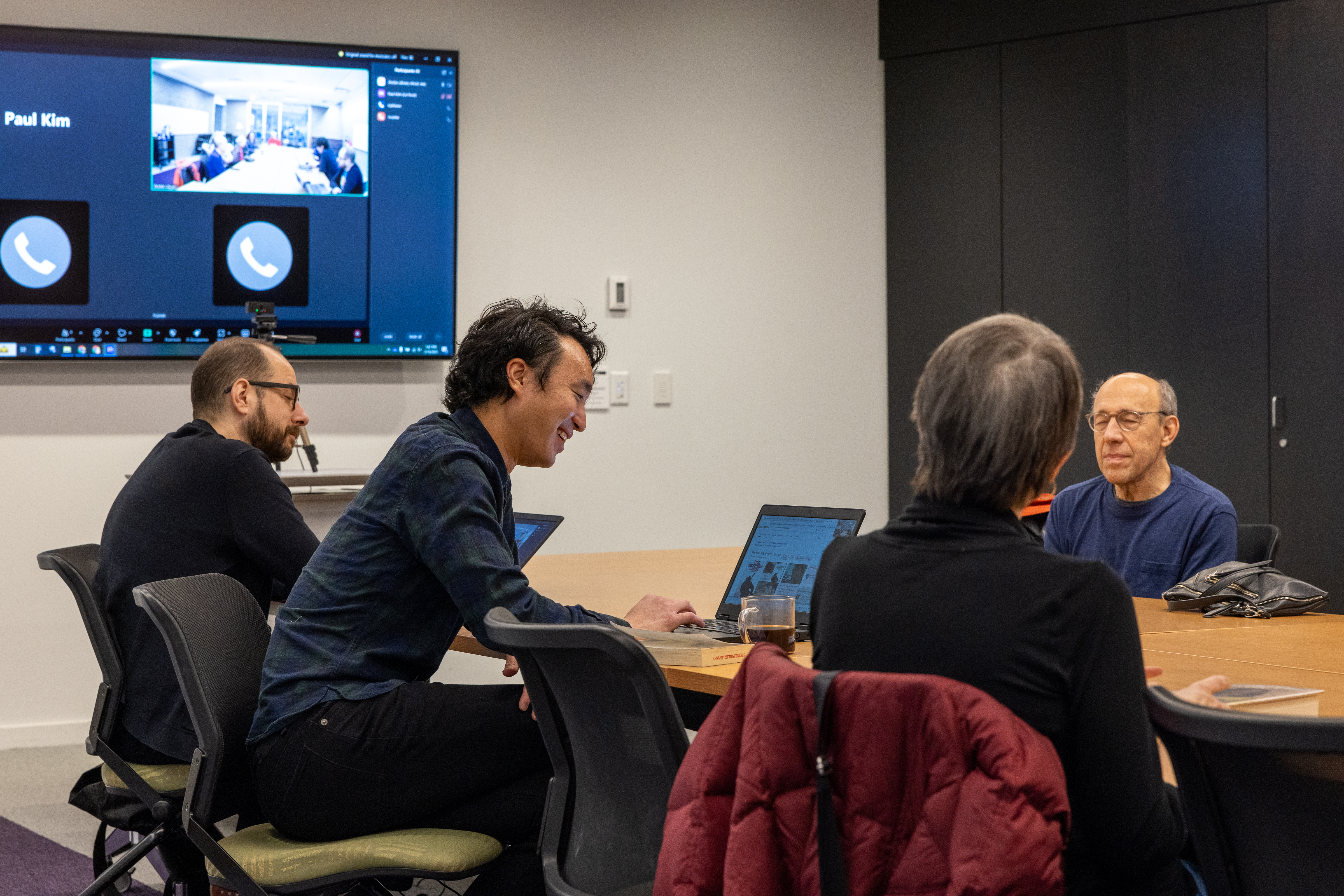 four people sitting across from each other at a table. One person has a laptop and there are zoom participants on a screen on the wall four people sitting across from each other at a table. One person has a laptop and there are zoom participants on a screen on the wall