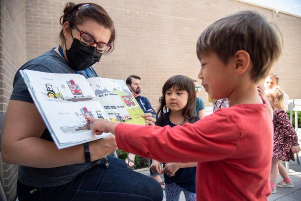 A staff member wearing glasses and a black mask holds a book with pictures of trucks as part of a storytime. A child with a red long-sleeve shirt is pointing at the book while another child with brown hair looks on.