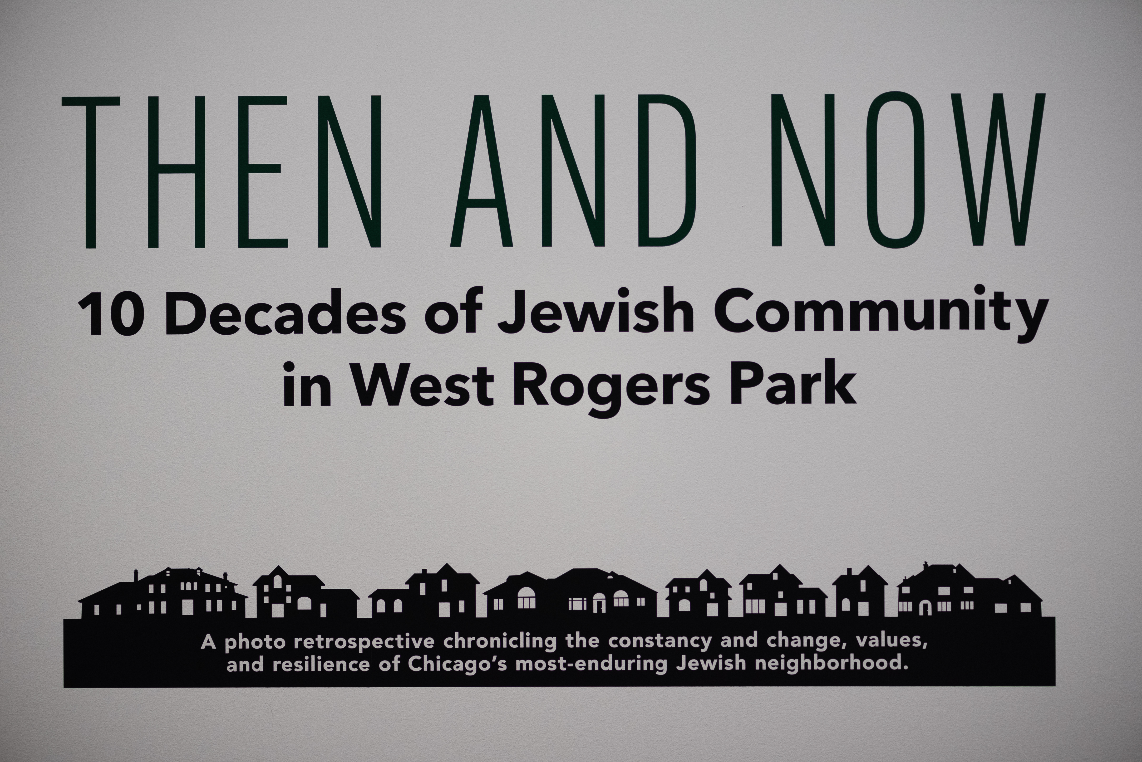 vinyl letters on a wall read "Then and Now: 10 Decades of Jewish Community in West Rogers Park" There is an illustration of a housing below vinyl letters on a wall read "Then and Now: 10 Decades of Jewish Community in West Rogers Park" There is an illustration of a housing below