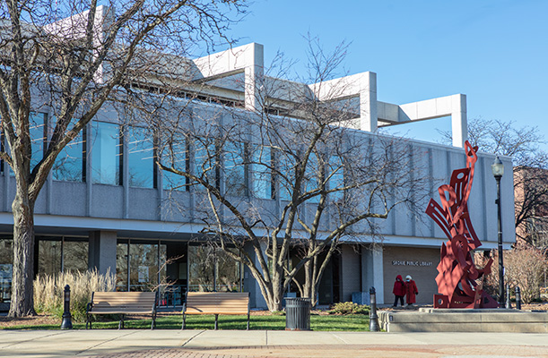 The east exterior of the Skokie Public Library on a sunny day, showing a large red sculpture outside, trees, and a row of second floor windows.