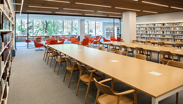 The current Reading Room in the Skokie Public Library, showing long wooden tables with wooden chairs, bookshelf-lined walls, and low orange chairs along a floor to ceiling wall of glass that looks out on the second floor of the library.