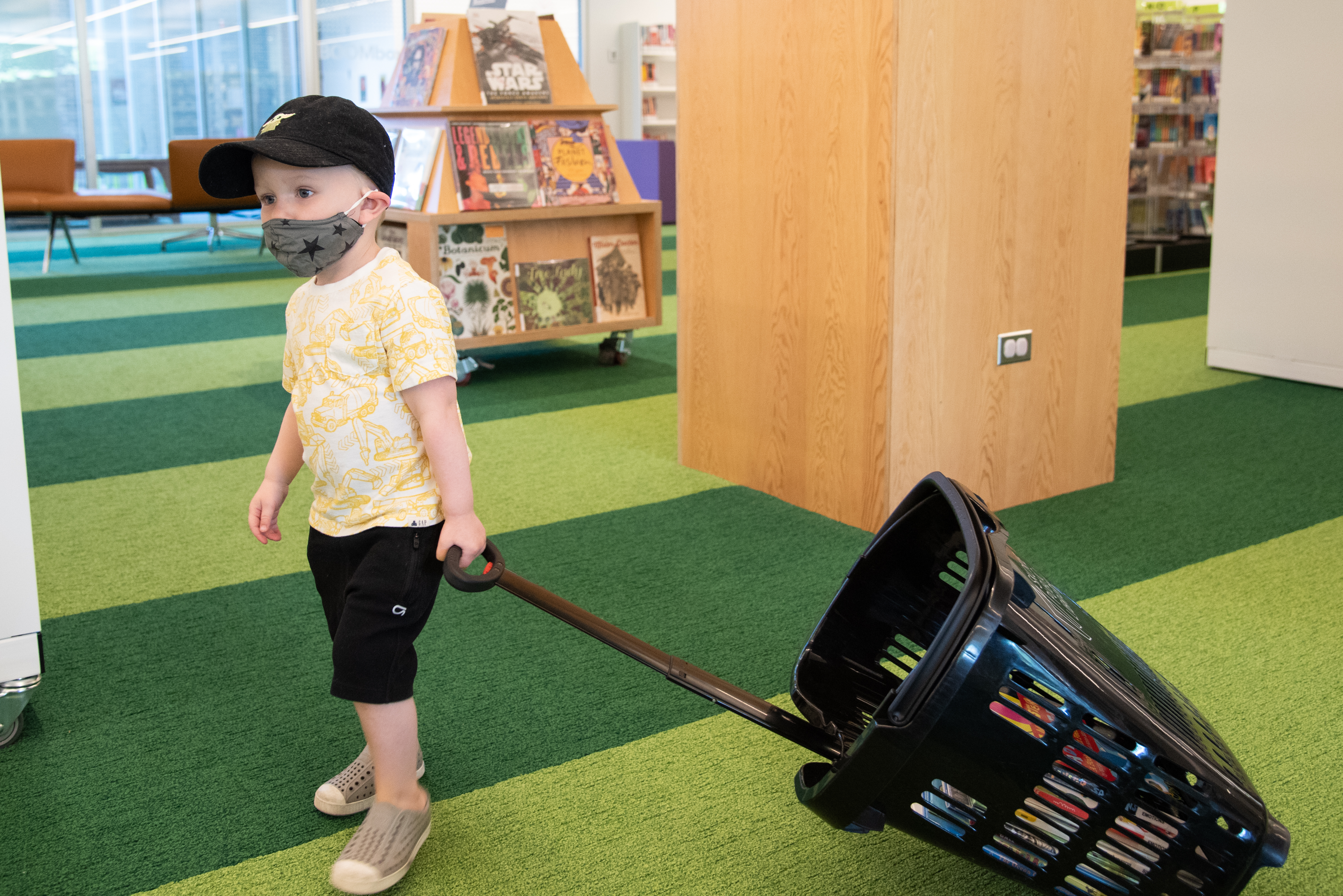 A child wearing a masks pulls a black wheeled book basket through the Kids area of the Skokie Public Library.