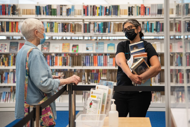 A patron in blue talks to a staff member in black, who is carrying a large stack of books. There is a large white bookshelf behind them.
