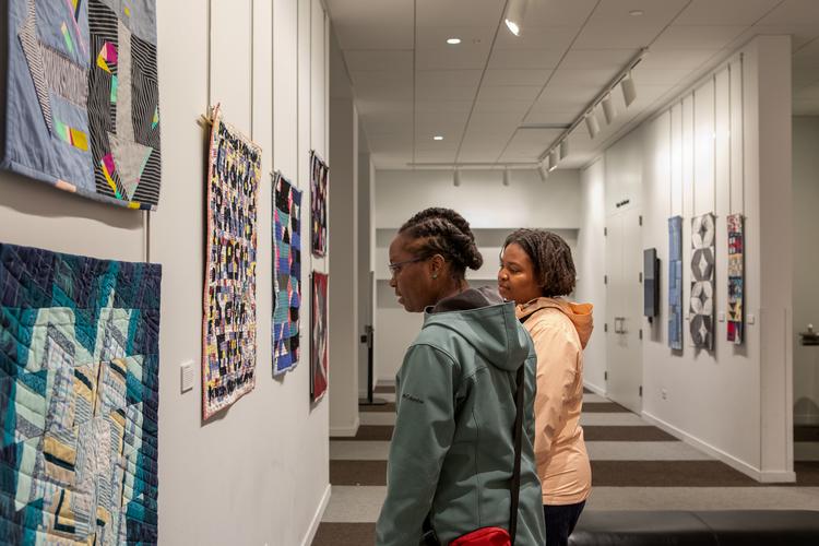 Two people look at small quilts displayed on a gallery wall