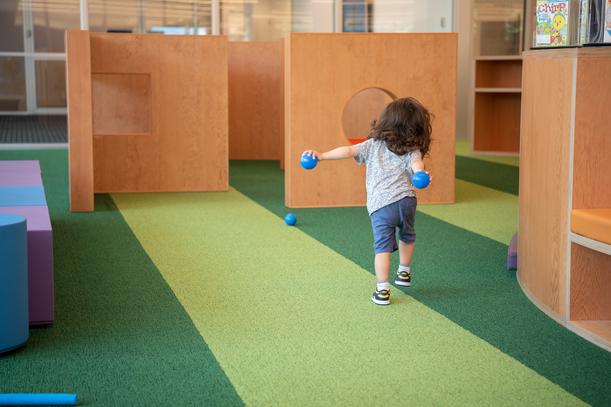 A young kid with their back to the camera marches through the Kids Room with two balls in their hands.