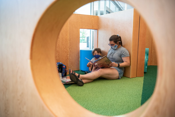 A woman and a child are seen through a round hole in a wooden wall. They are sitting on the green floor of the Kids Room, reading together.