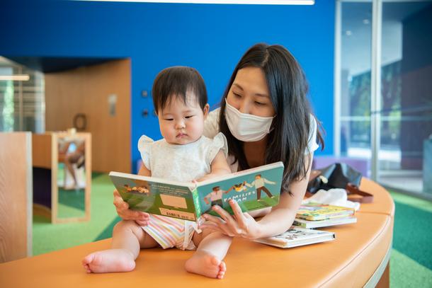 A masked woman reads a book to her baby daughter, wearing white, who is seated on an orange bench. The book has a family illustrated on the front cover.