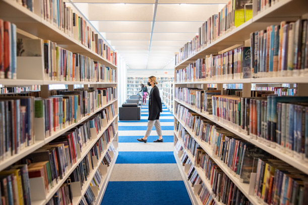 Two long white bookshelves stretch to the center of the frame, where a staff member is walking by on the blue and gray striped carpet.