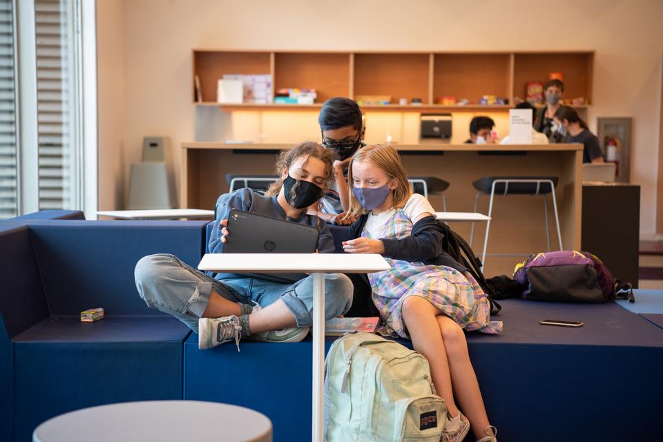 Three teens lean towards each other while looking at the same laptop in the common area of the Teen Room.