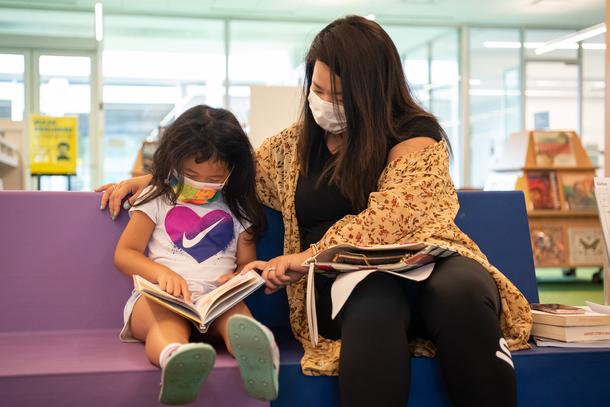 A woman wearing a brown sweater sits next to her daughter, who holds a book in her lap. The woman points to something in the book as the girl reads.
