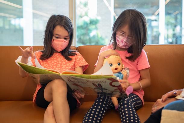 Two young girls wearing pink shirts and masks read a book together on a brown bench. One girl is holding a baby doll.