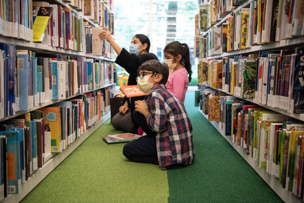 A woman and two children sit on the green floor of the Kids Room, browsing for books between two tall shelves. The woman is reaching for a book.