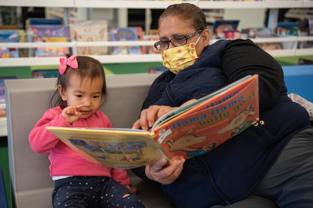 A masked woman sits next to a child, reading a book to her. The woman holds her hand to the page and the girl points to the page as well. The girl is wearing a pink shirt and a pink bow and the woman is wearing a yellow mask.