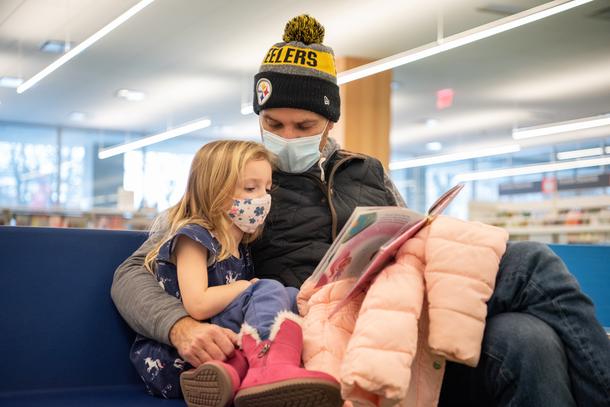 A father wearing a black and yellow beanie with a pom-pom reads to his daughter, who is curled up to his side.