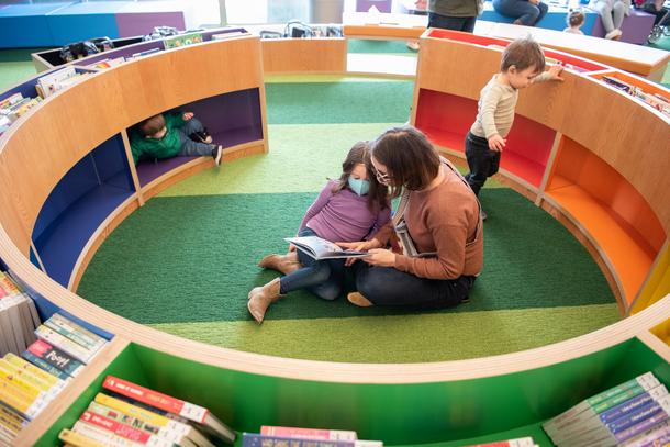 A woman reads to her daughter in the center of a circular shelf with multiple colors including blue, purple, red, orange and green. Her two other boys are playing around the shelves.
