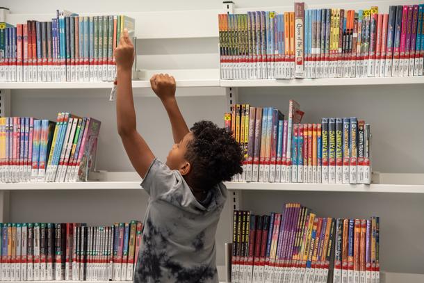 A child reaching high above their head to pick a book off of the top shelf. The shelves are white and the books are colorful.