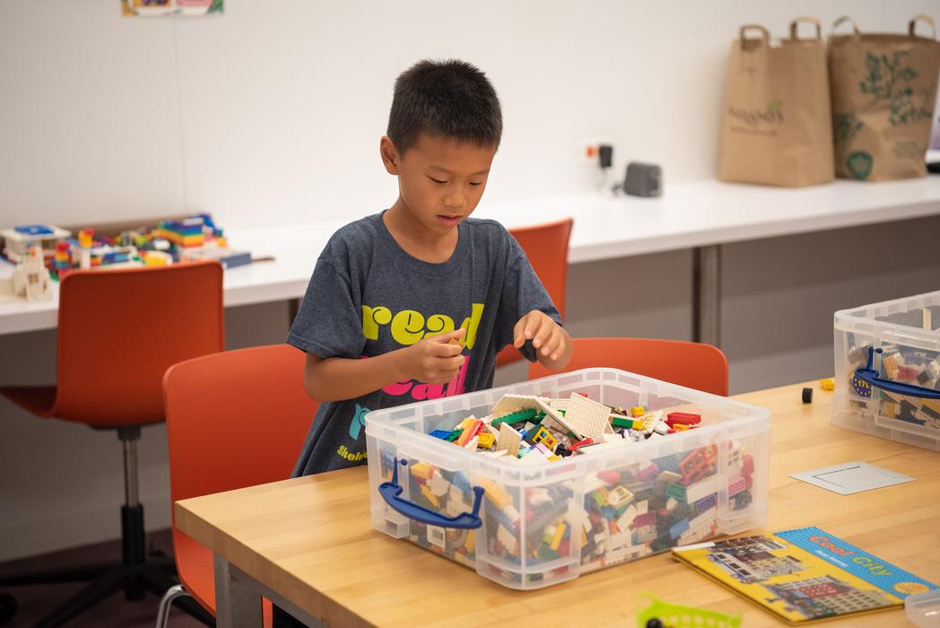 A boy with short, dark hair wearing a grey t-shirt with the word "Read" in yellow letters picks out Legos from a bin